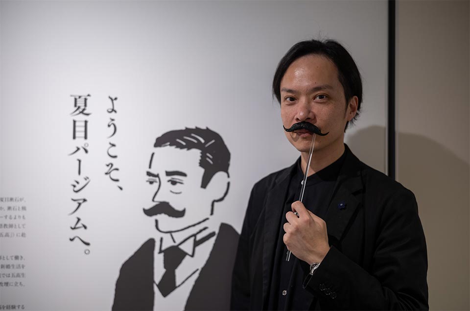 漱石のトレードマークの口髭(ひげ)を付けてポーズを取る佐藤さん。夏目パージアムのオーナーであると同時に建築家でもあります。