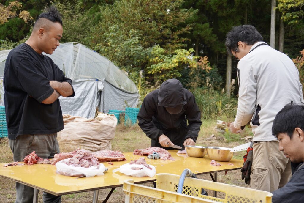 屋外の机で包丁を手に肉をさばく研修生たちと見守る鳥越さん