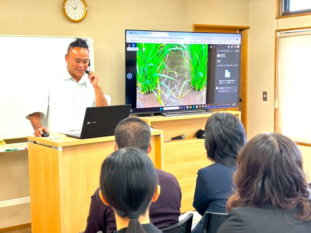 会議室の演台でパソコンとヘッドセット、モニターを使って講演する鳥越さん。画面には水田の稲が映し出されている
